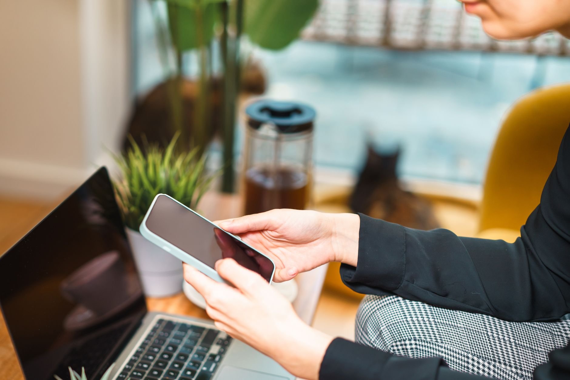 Woman working remotely from home, using a smartphone and laptop at a cosy desk.