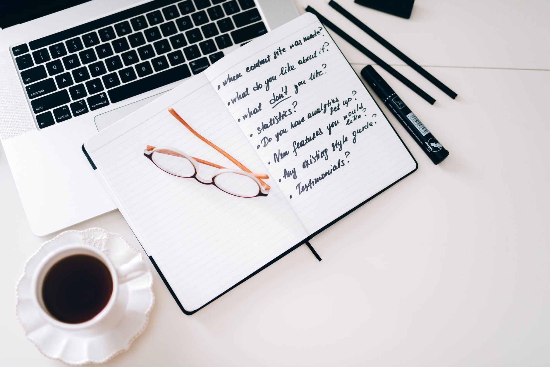 A stylish workspace featuring a laptop, notebook with notes, glasses, and a coffee cup.